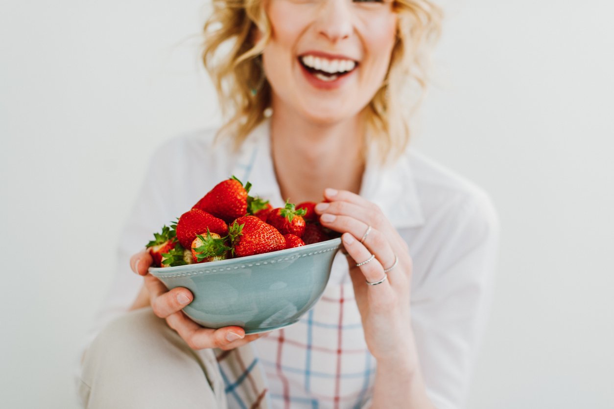 Oberkörper und untere Gesichtshälfte einer Frau mit blonden Locken. Sie trägt ein kariertes Hemd und lacht. Dabei hält sie mit beiden Händen eine Schüssel mit großen, roten Erdbeeren.